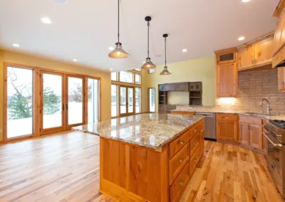 Modern kitchen interior with wooden cabinets and granite countertops, featuring pendant lighting and large windows with a view outside.