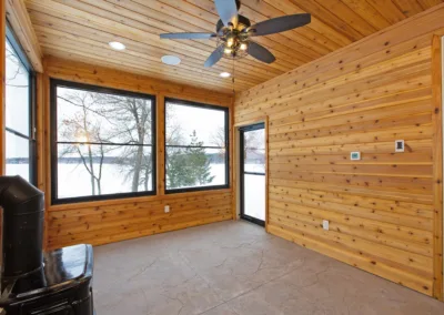 A bright, empty room with wooden walls, a ceiling fan, and large windows showcasing a snowy landscape outside.