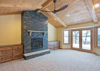 A spacious room featuring a stone fireplace, wooden beamed ceiling, and large windows with a view of snowy landscape outside.