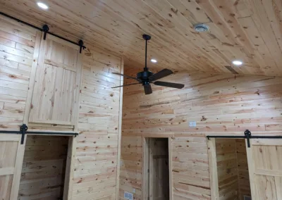 Interior of a wooden cabin with paneled walls and ceiling, featuring a black ceiling fan and built-in cabinets with iron hardware.