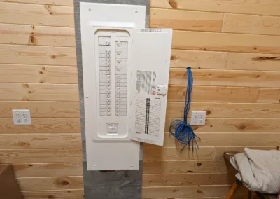 Electrical circuit breaker panel with open door mounted on wooden wall and unfinished networking cables to the side.