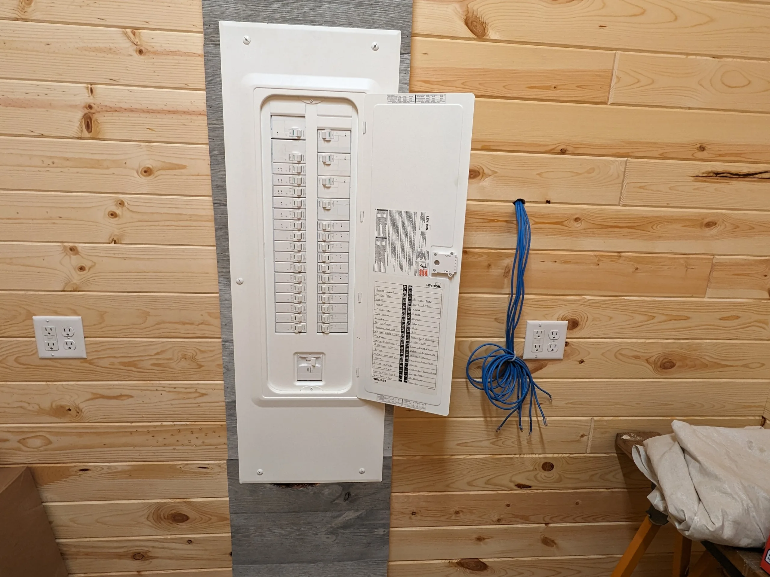 Electrical circuit breaker panel with open door mounted on wooden wall and unfinished networking cables to the side.