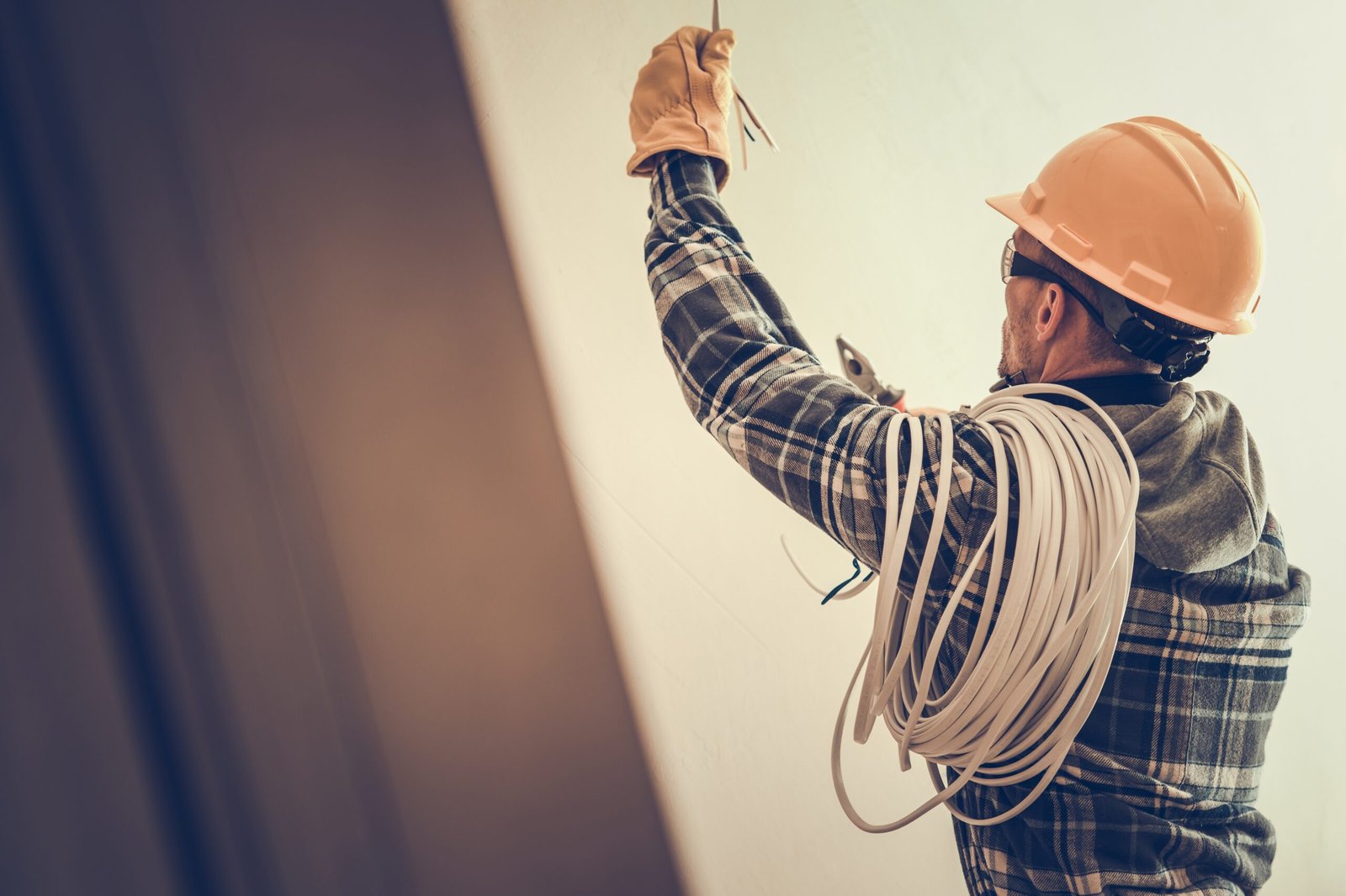 An electrician in a hard hat and gloves installs wiring on a wall, carrying a bundle of cables on his shoulder.