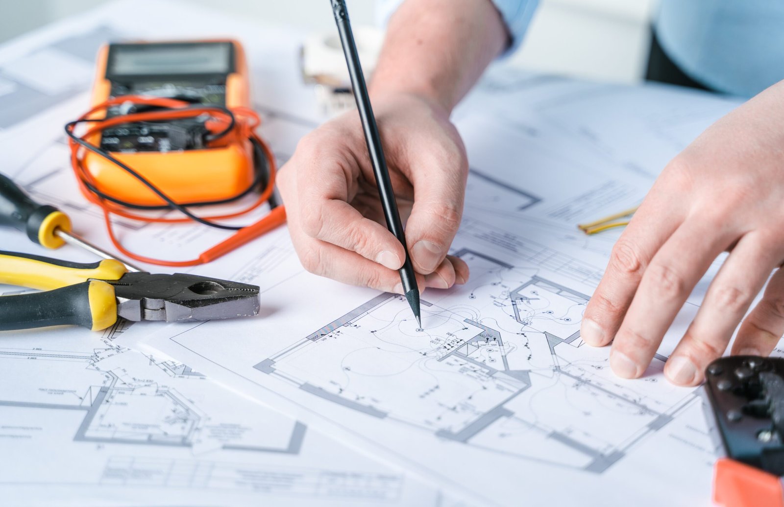 Two hands, one holding a pencil, marking on architectural blueprints with various tools like a multimeter and pliers nearby.