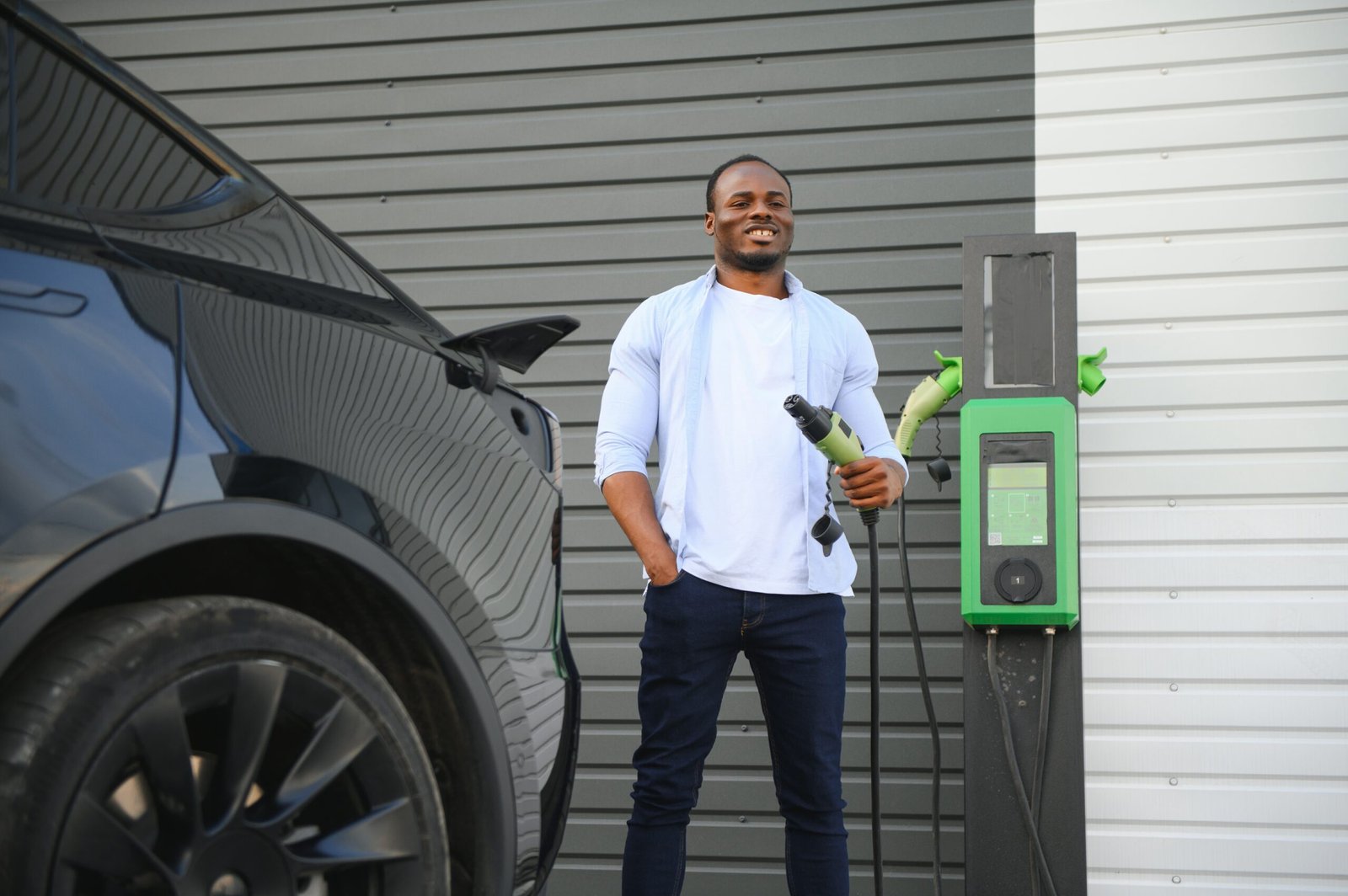 A smiling man holding a charging plug next to an electric car at a charging station.