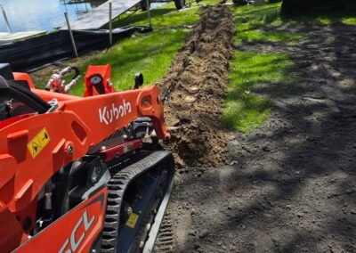 A Kubota SCL 1000 track loader sits on a dirt path beside a freshly dug trench near a lakeside dock, with people and trees in the background.