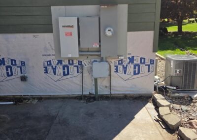 Electrical panels and a Generac unit are mounted on the side of a house under a window, with exposed insulation and an air conditioning unit nearby.