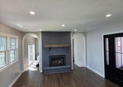 A living room with dark wood floors, light-colored walls, arched doorways, a gray brick fireplace with a wooden mantel, and large windows letting in natural light.