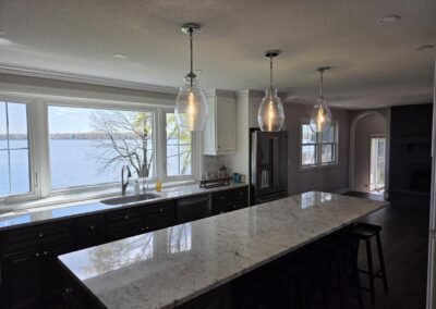 Modern kitchen with a large island, three pendant lights, dark cabinets, and a view of a lake through wide windows.