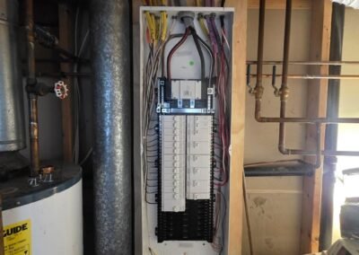Electrical panel with multiple circuit breakers and organized wiring mounted on a wooden wall in a utility room; pipes and a water heater are visible nearby.