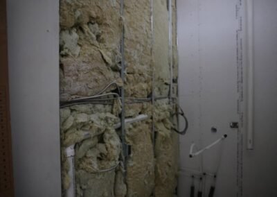 Exposed wall with insulation foam, electrical wiring, and plumbing pipes visible; unfinished drywall surrounds the area.