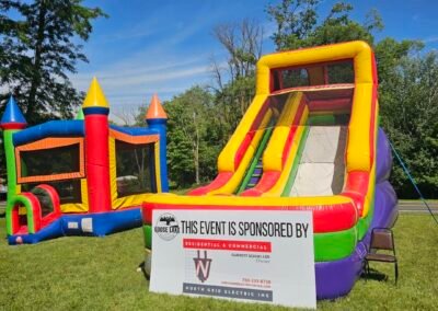 Two inflatable bounce houses, one with a slide, stand on grass beside a sponsorship sign for North Grid Electric Inc. Trees and blue sky are visible in the background.